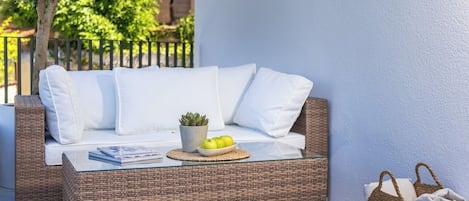 Un canapé en osier avec des coussins blancs et une table basse en verre, placés sur une terrasse carrelée, avec un panier en osier et une plante en pot à proximité.