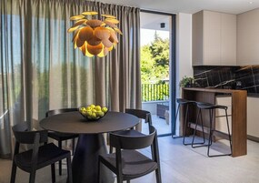 A modern dining area with a round table, black chairs, and a bowl of lemons. There is a kitchen with a black countertop and a hanging light fixture.
