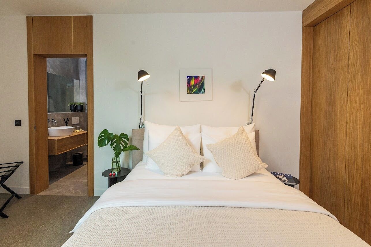 A bedroom with a bed, bedside lamps, a nightstand, a potted plant, and a bathroom visible through an open door.