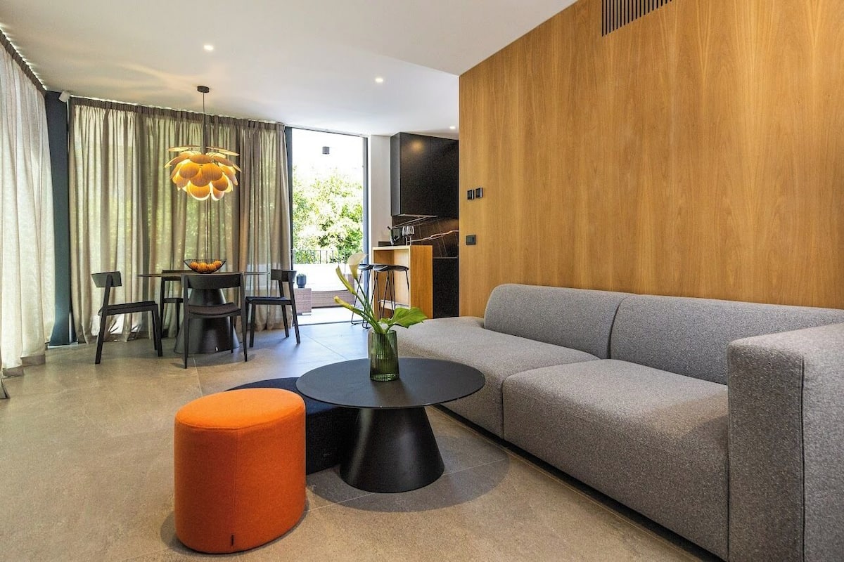 A modern living room with a grey sofa, a round black coffee table, and an orange ottoman. There is a dining area with a table and chairs, and a large window with curtains.