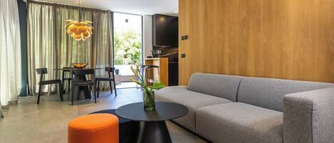 A modern living room with a grey sofa, a round black coffee table, and an orange ottoman. There is a dining area with a table and chairs, and a large window with curtains.