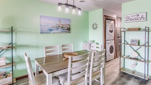 A dining area with a wooden table and white chairs, a painting of a lighthouse, a washing machine, and a shelf with decorative items. - Walk to Ocean: Condo w/ 3 Pools in Myrtle Beach (Myrtle Beach)