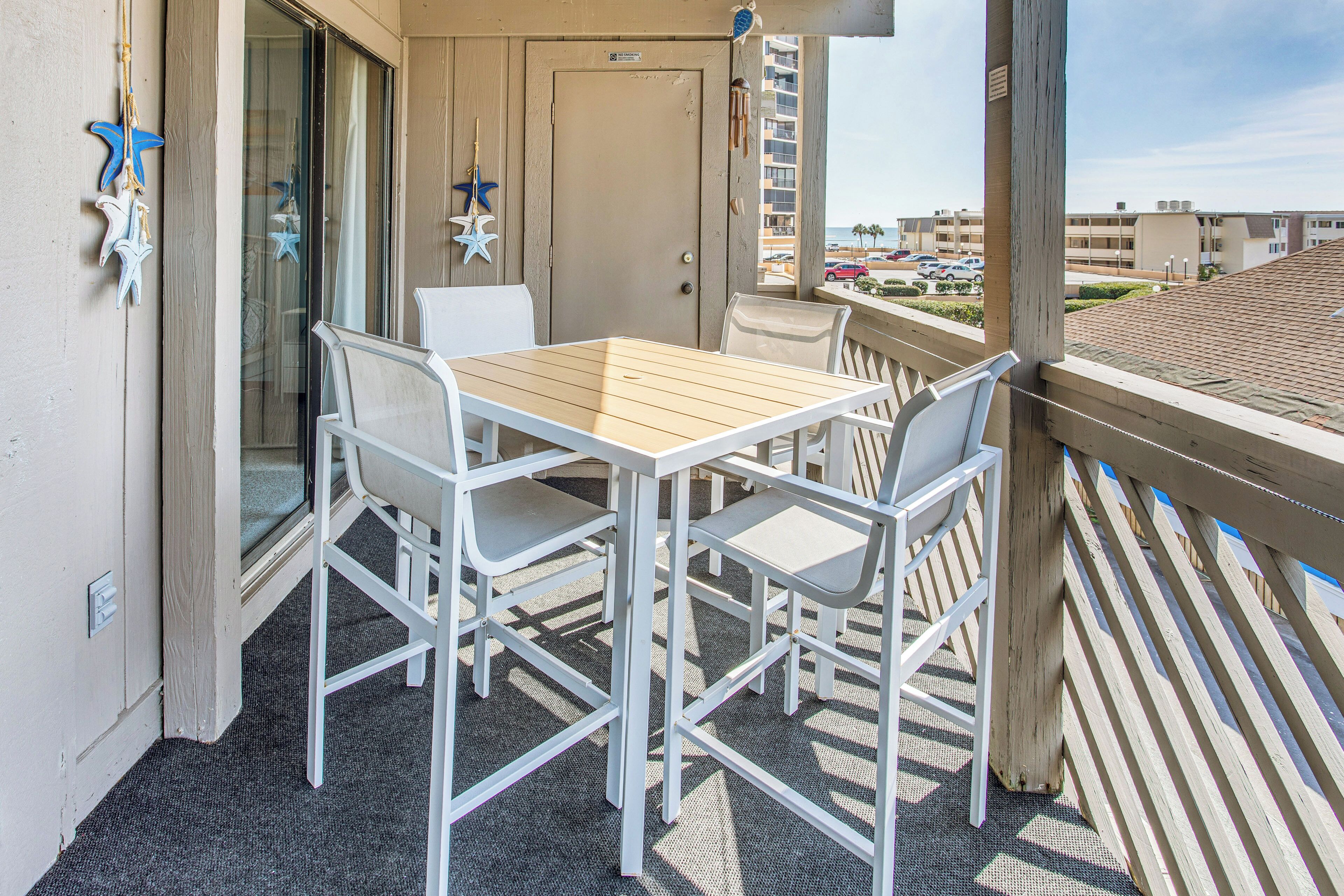 A balcony with a wooden table and white chairs, a door, and a view of the beach and other buildings.