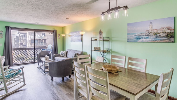 A dining area with a wooden table and chairs, a sofa, and a view of the balcony. - Walk to Ocean: Condo w/ 3 Pools in Myrtle Beach (Myrtle Beach)