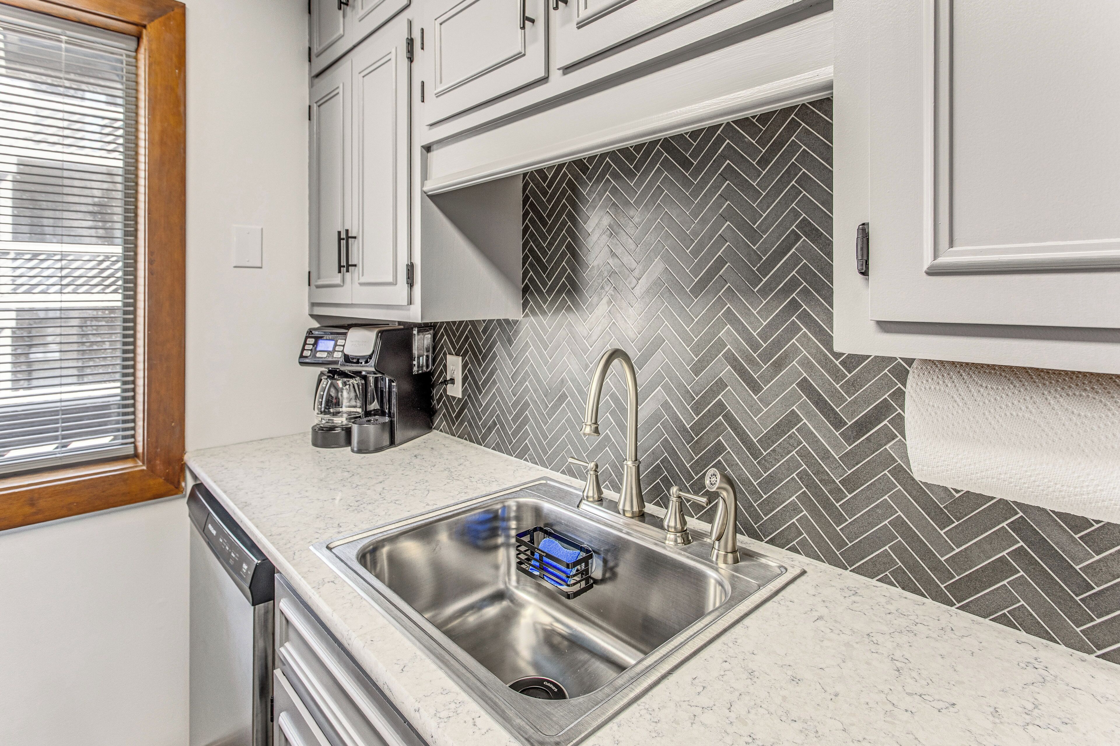 A kitchen with a stainless steel sink, white cabinets, a herringbone-patterned backsplash, and a window with blinds.