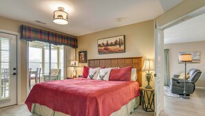 A spacious bedroom with a large bed, a view of the outdoors through a sliding door, and a cozy reading nook with a chair and lamp. - On Thousand Hills Golf Course: Condo w/ Sunroom (Branson)
