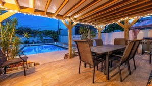 Un patio exterior con piscina, pérgola de madera, luces colgantes, mobiliario de mimbre y una parrilla.