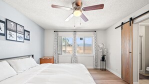 A bedroom with a bed, a ceiling fan, a window with curtains, a wooden door, and framed pictures on the wall.