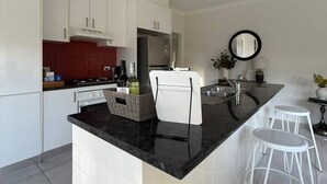 Cocina moderna con armarios blancos, encimera negra y salpicadero rojo. Hay dos taburetes blancos tipo bar y un espejo redondo en la pared.