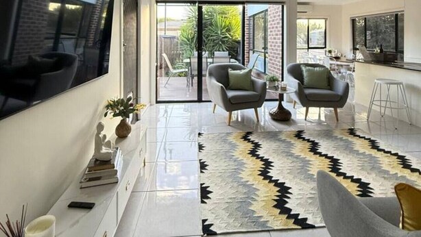 Una sala de estar moderna con un televisor grande, dos sillones, una alfombra y una cocina visible a través de puertas de cristal.