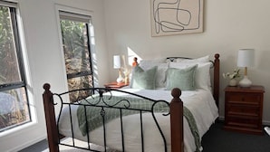 Un dormitorio con estructura de cama metálica, una mesita de noche con lámpara y ventana con vista a follaje.