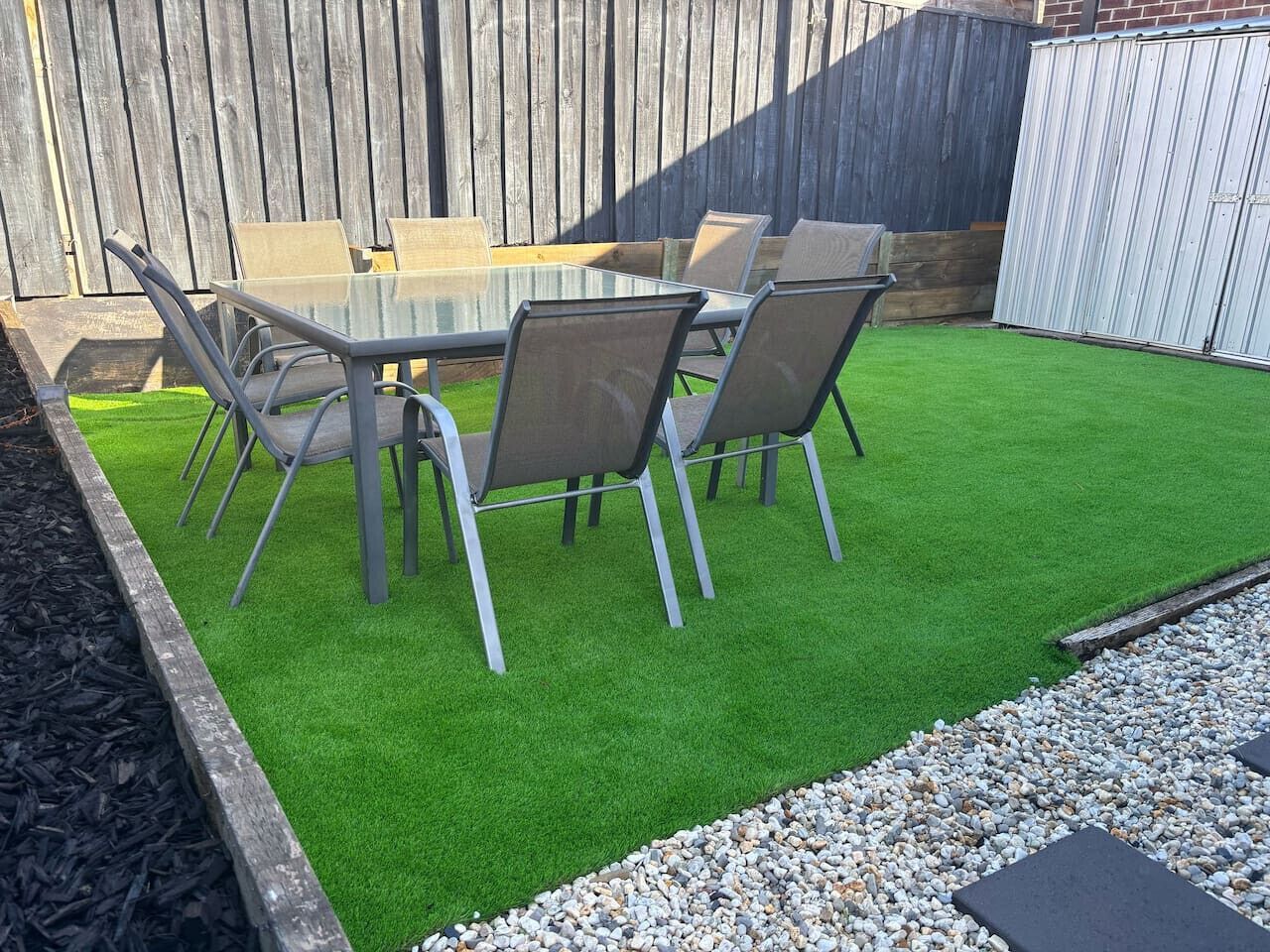 Une terrasse avec une table et des chaises en verre, entourée de gazon synthétique et d’une clôture en bois.
