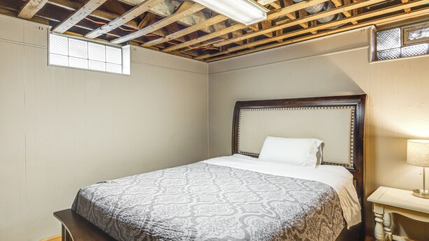 Un dormitorio con techo de madera, una cama con colcha estampada, una mesita de noche con lámpara y un ventanal con celosía decorativa.