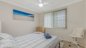 Une chambre à coucher avec un lit, une chaise, une fenêtre avec des stores, un ventilateur de plafond et un tableau accroché au mur.
