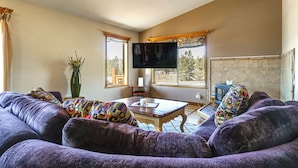 A living room with a sofa set, a coffee table, a flat-screen TV, and large windows offering a view of trees. - Mtn-view Getaway w/ On-site Trails in Fairplay! (Fairplay)