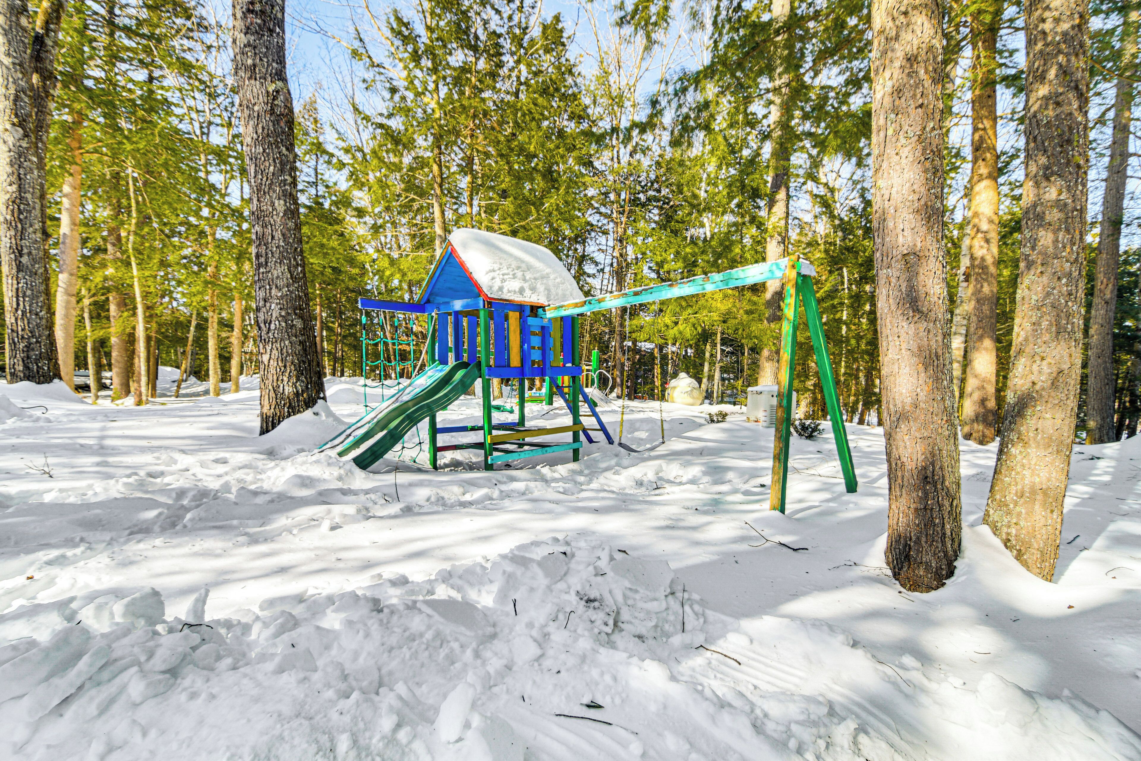 A snow-covered playground with a slide and swings, surrounded by tall trees.
