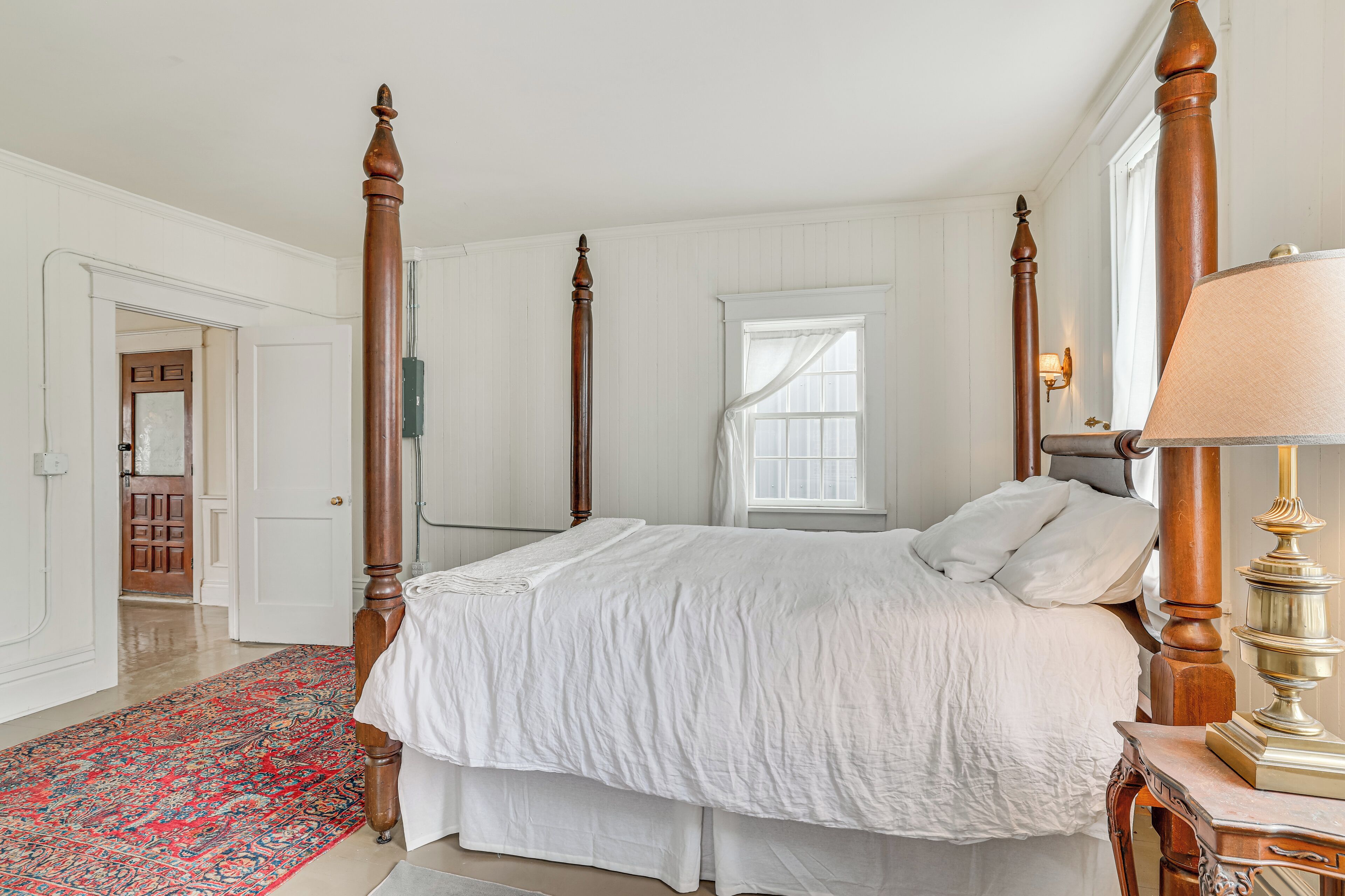 A four-poster bed with white bedding, a red patterned rug, and a wooden nightstand with a lamp.