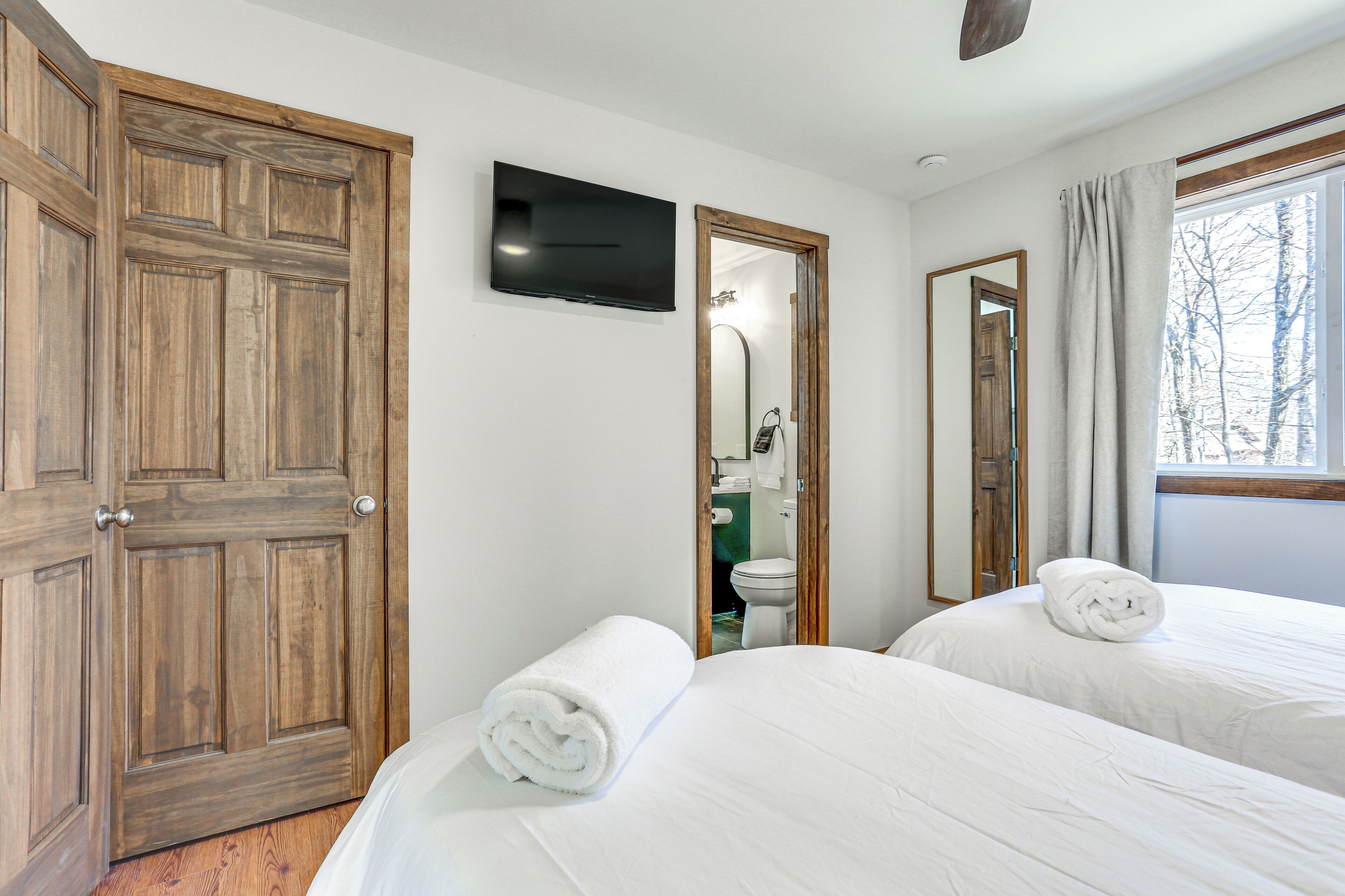 Hotelkamer met twee bedden, een televisie, houten deuren, een zichtbare badkamer via een open deur en een raam met gordijnen.