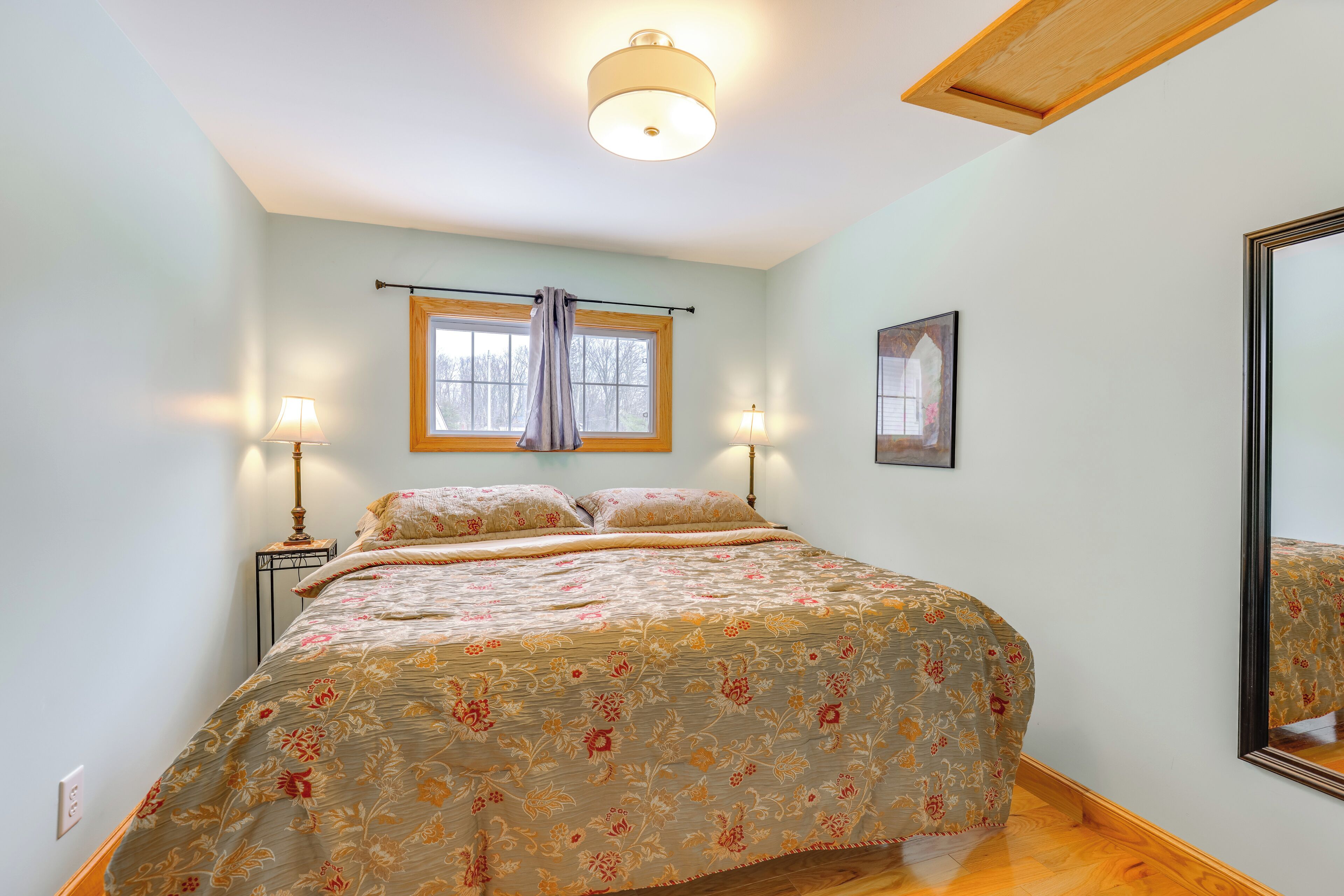 A bedroom with a bed, two bedside lamps, a window with curtains, a framed picture on the wall, and a ceiling light.