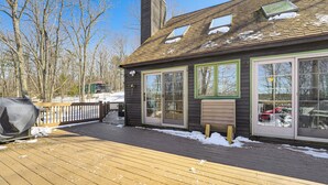 Eine Holzdiele mit Schnee, ein überdachter Grill und ein Haus mit großen Fenstern und Schornstein.