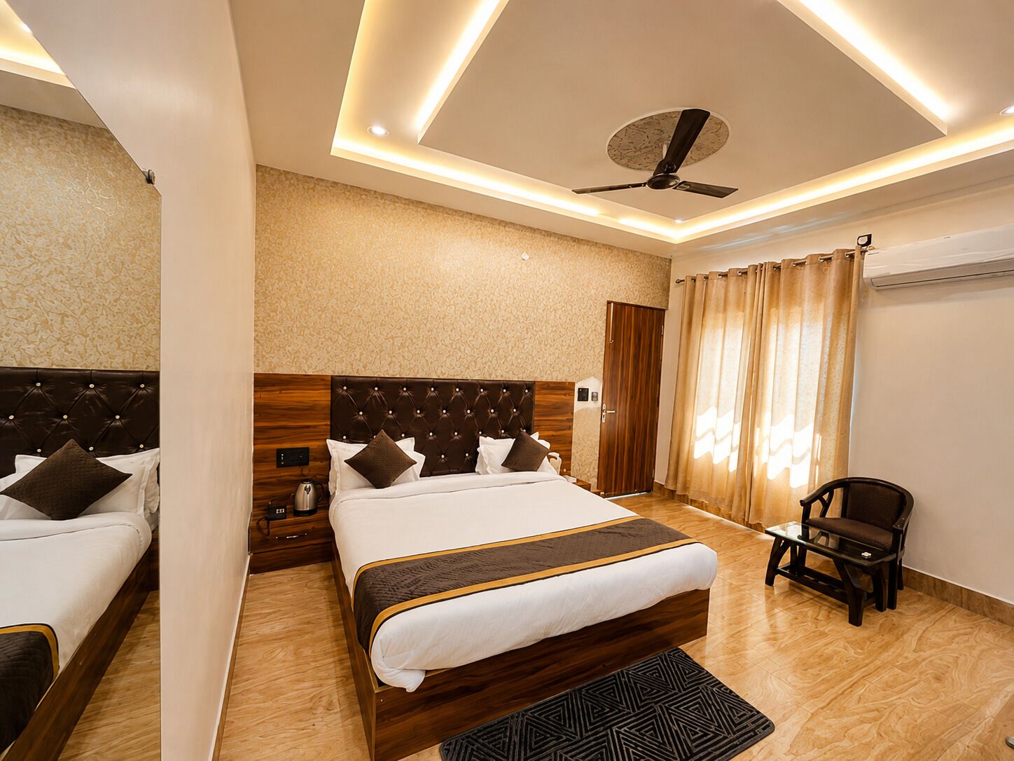 A hotel room with a large bed, a smaller bed, a desk, a chair, a ceiling fan, and an air conditioning unit.
