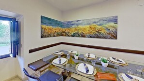 A dining area with a glass table set for six, a large painting above the table, and a window on the left.