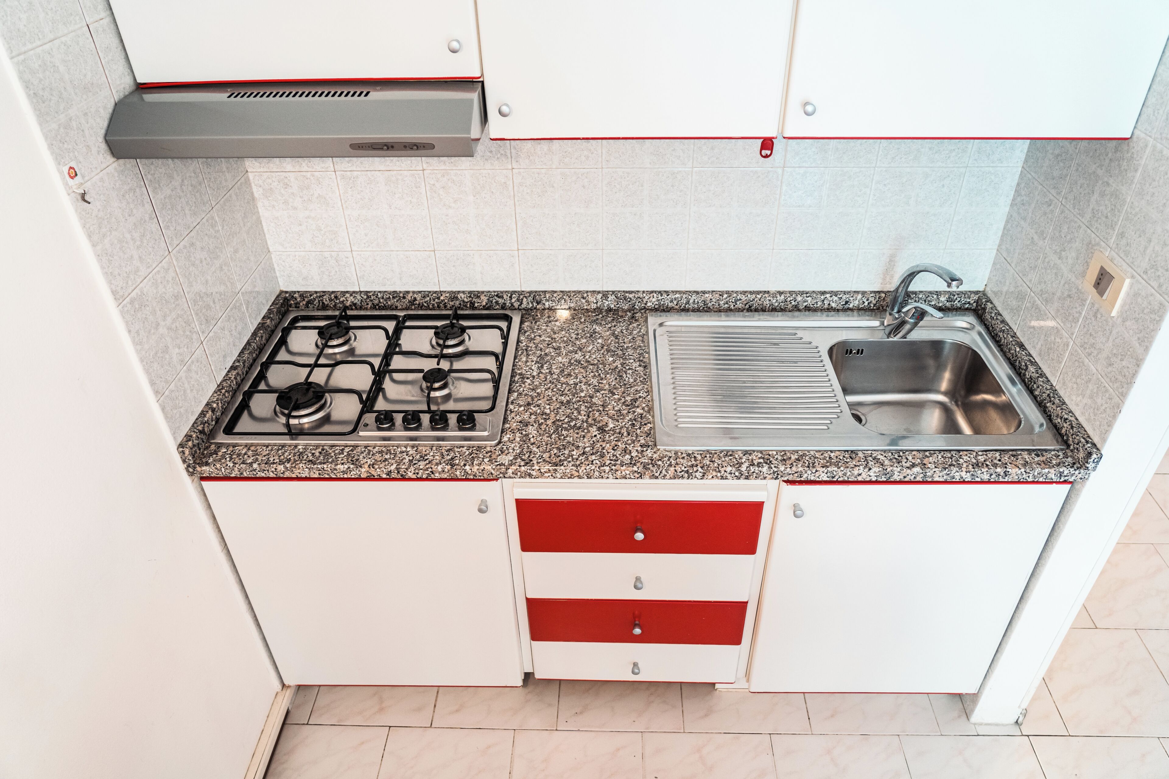 A compact kitchen with a gas stove, stainless steel sink, and white cabinets with red handles.
