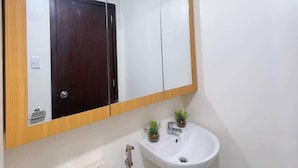 A bathroom with a large mirror, a white sink, a small potted plant, and a towel dispenser. - Cozy Condo in Mandaluyong City, Metro Manila (Mandaluyong)