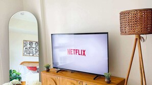 A living room with a television displaying 'NETFLIX' on the screen, a wooden cabinet below, and a woven lamp to the right. - Cozy Condo in Mandaluyong City, Metro Manila (Mandaluyong)