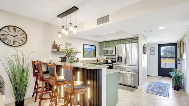 A modern kitchen with a bar area, stainless steel appliances, and a large clock on the wall. - The Gator Escape -Poolside Condo walk to Tiki Bar (Clewiston)
