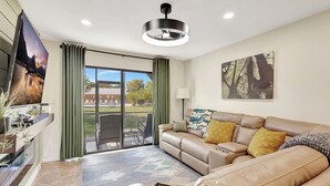 A modern living room with a leather sofa, a flat-screen TV, a large window with curtains, and a ceiling light fixture. - The Gator Escape -Poolside Condo walk to Tiki Bar (Clewiston)