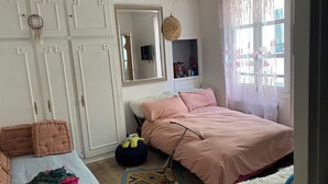 Une chambre à coucher comprenant un lit, une armoire, un miroir, une fenêtre avec des rideaux à motifs floraux et une petite étagère ornée d’objets décoratifs.