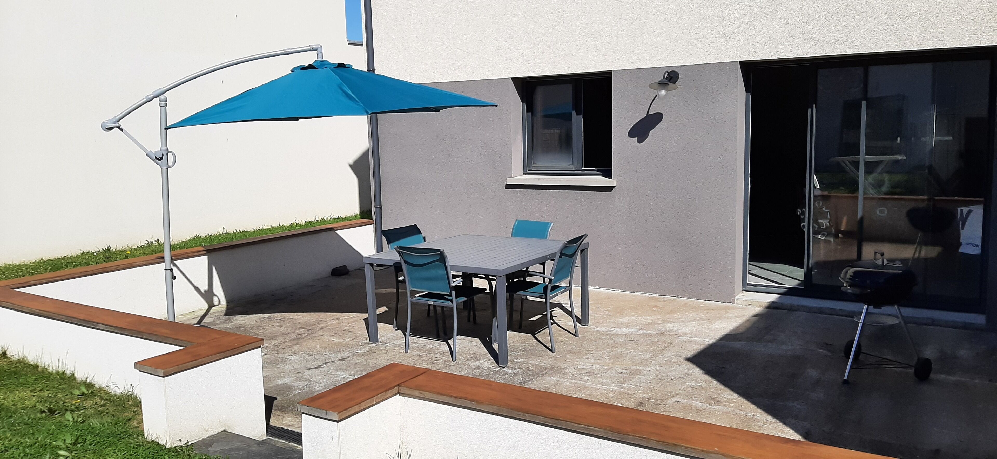 Une terrasse extérieure avec une table et des chaises, un parasol bleu et une porte vitrée donnant sur un espace abrité.