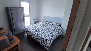 Une petite pièce bien éclairée, équipée d’un lit, d’un bureau avec un ordinateur et une imprimante, d’une armoire et d’une fenêtre laissant entrer la lumière naturelle.