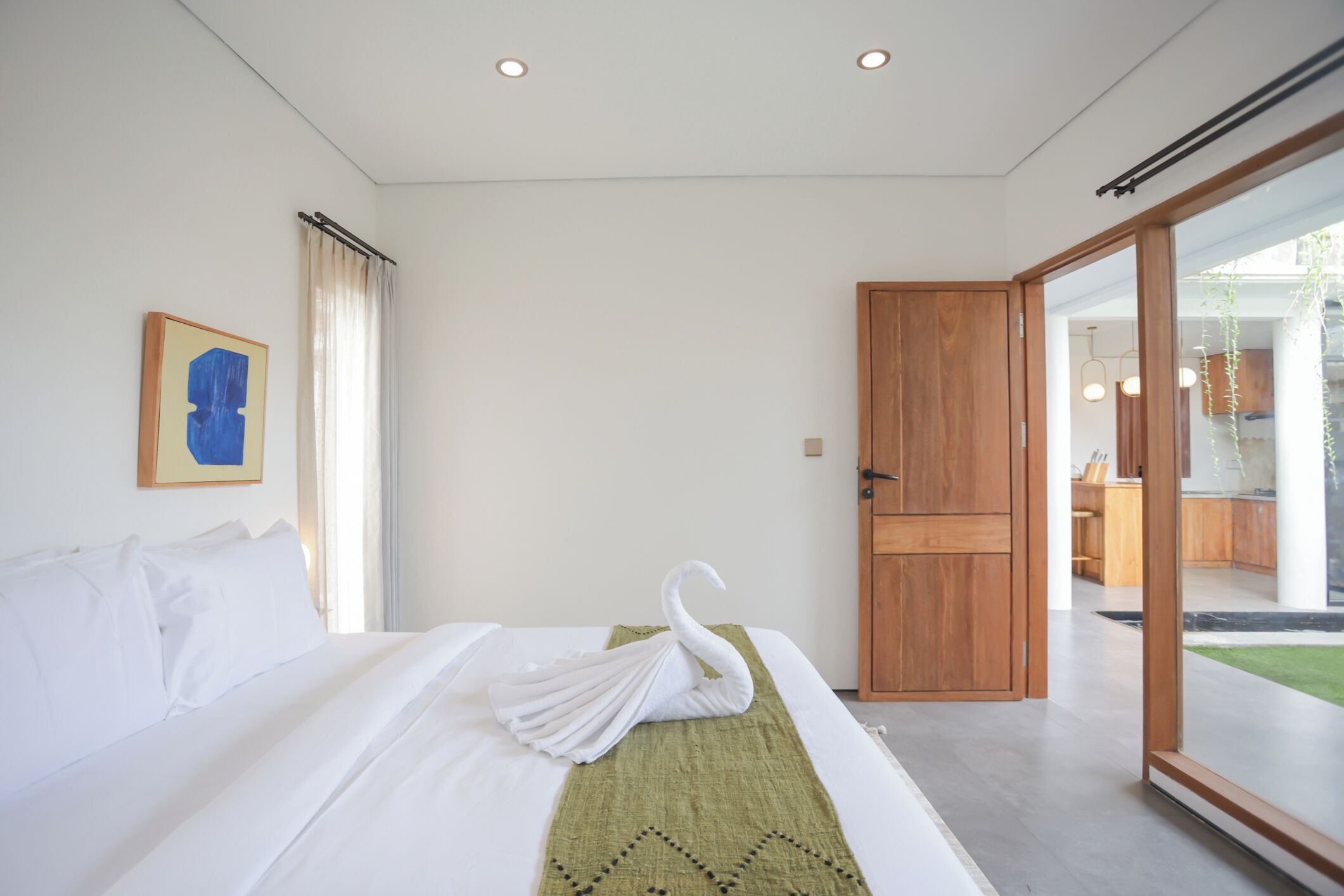 A modern hotel room with a neatly made bed, a wooden door, and a view of the outdoor area.