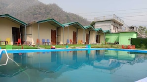 A pool area with a clear blue pool, surrounded by a row of single-story buildings with green roofs and wooden doors. - Sunrise Resort (Shivpuri)