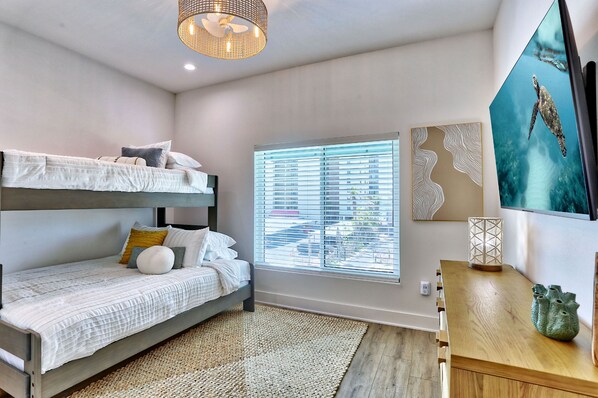 A bedroom with a bunk bed, a desk, a window with blinds, and a wall-mounted TV.