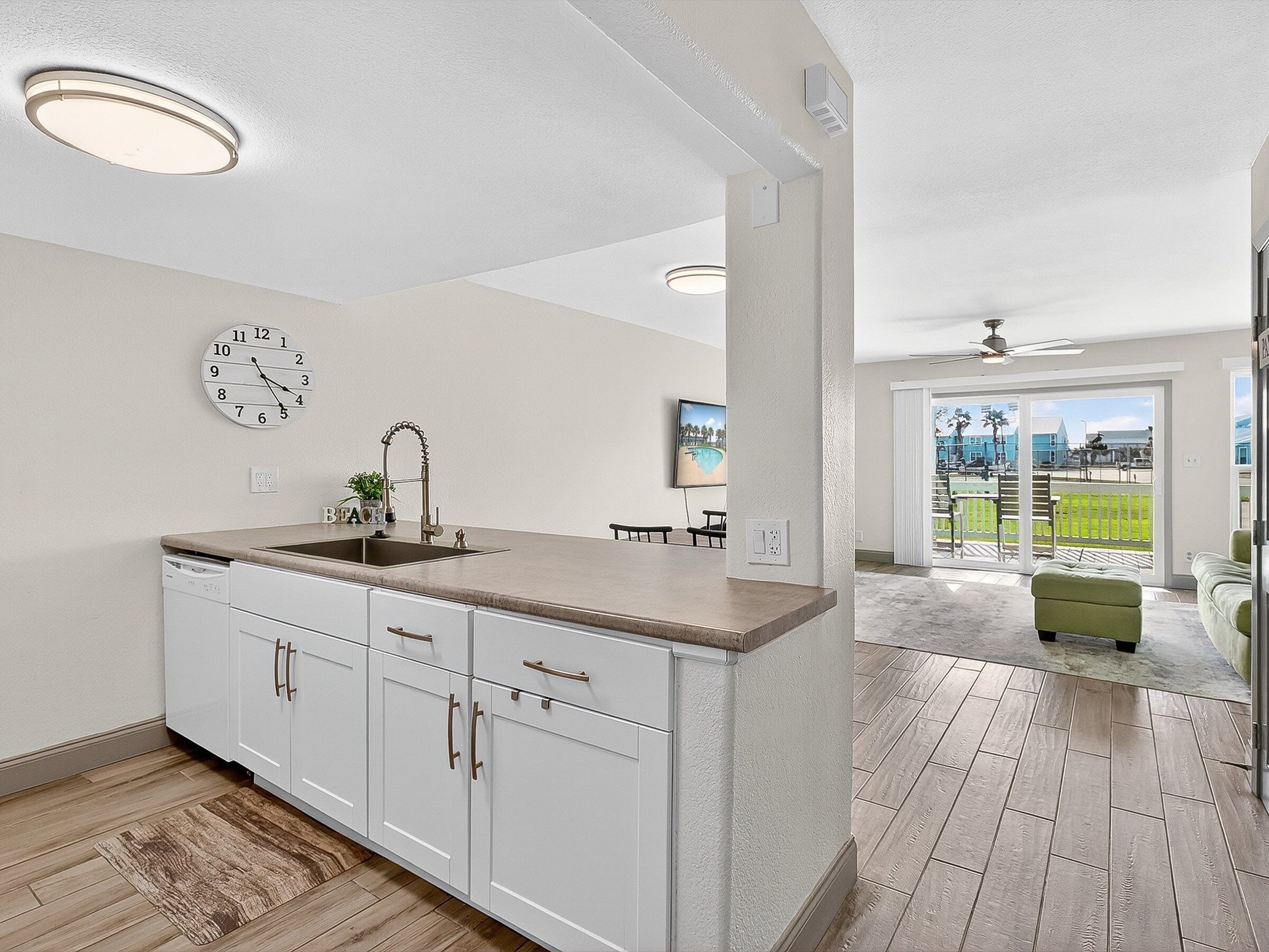 Una cocina con gabinetes blancos, un fregadero y un reloj en la pared. Hay una zona de comedor con una mesa y sillas, y vistas a un paisaje verde a través de una puerta de cristal.
