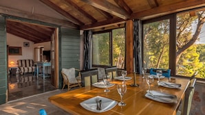 A dining area with a wooden table set for six, large windows with a view of trees, and a cozy seating area with a sofa and chairs. - Hidden Heaven Private Resort - Heated Pool (Milna)