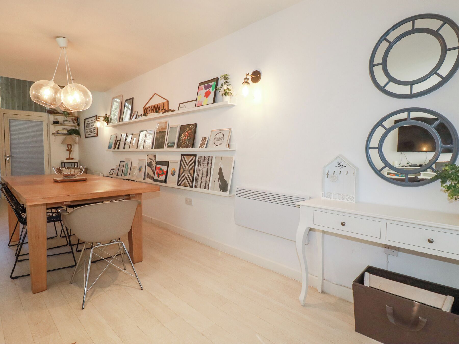 Un coin repas avec une table et des chaises en bois, une console blanche avec des tiroirs et des étagères murales ornées de tableaux encadrés.