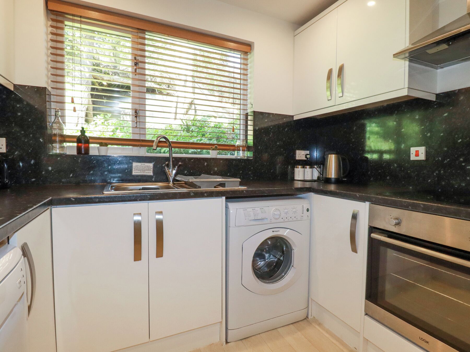 A modern kitchen with a built-in washer and dryer, a stainless steel oven, and a window with blinds.