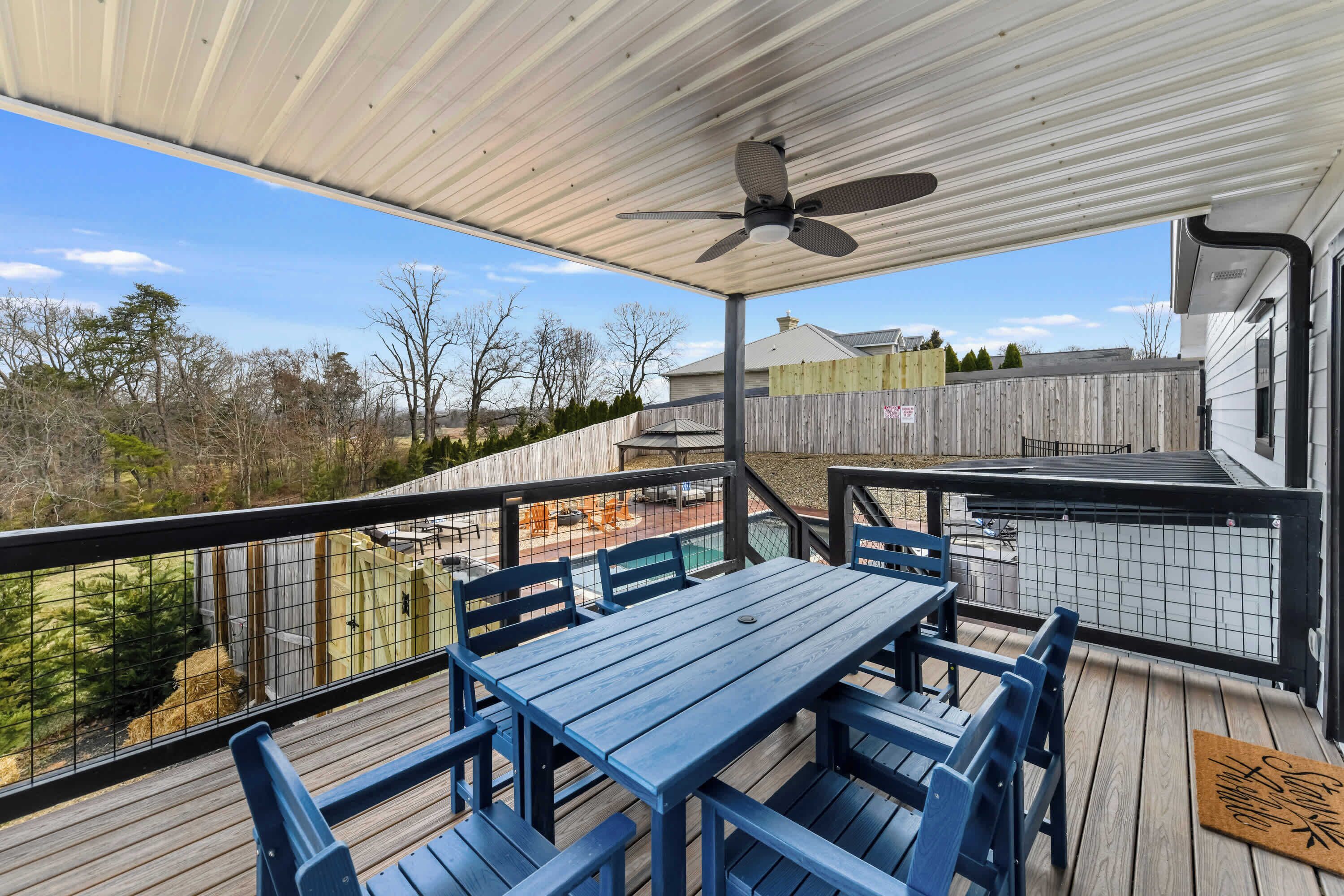 A covered outdoor deck with a table and chairs, a ceiling fan, and a view of a backyard with a pool and a wooden fence.