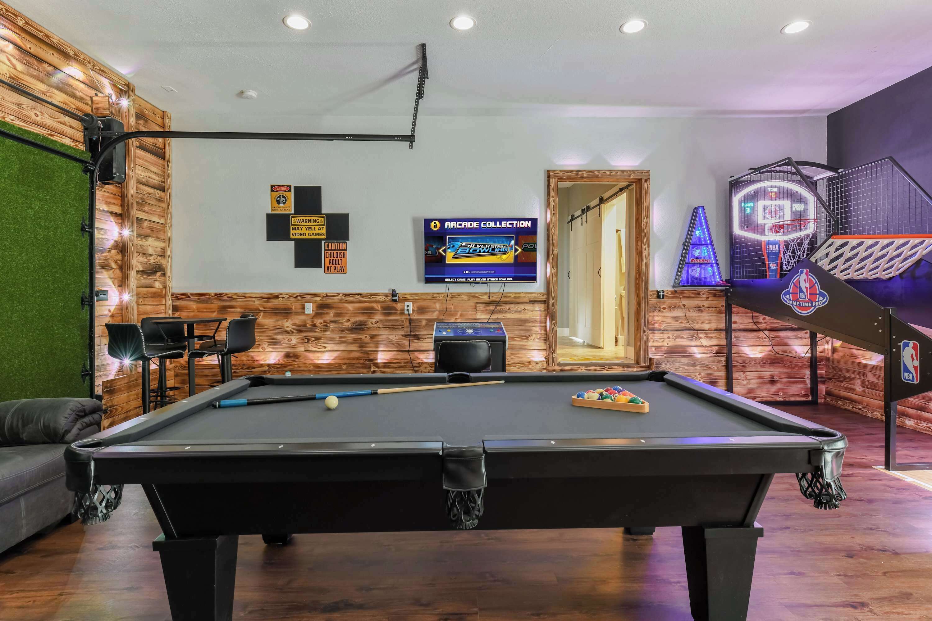 Ein Billardtisch mit einer weißen Billenkugel und einem Satz Billardstöcke, ein Fernseher, der 'Arcade Collection' anzeigt, ein Basketballkorb und eine Holzwand mit einer grünen Akzentwand.
