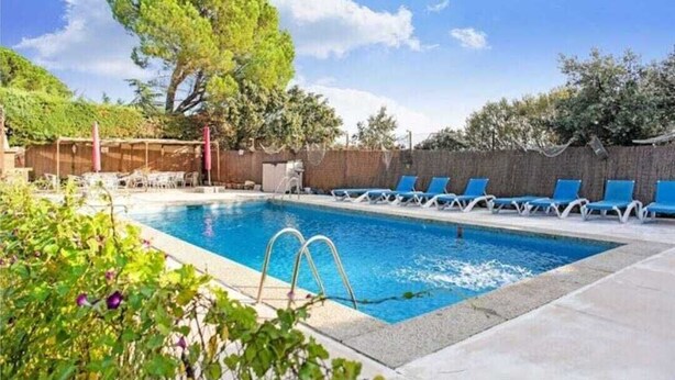 Une piscine rectangulaire à l’eau d’un bleu limpide, entourée d’un petit muret en briques. Plusieurs chaises longues sont alignées le long du muret, et une structure en bois formant une pergola est visible au fond.