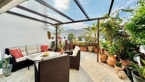 A covered patio with a sofa and table, surrounded by potted plants and a clear sky view.
