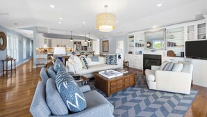 A spacious living room with a sofa, coffee table, and fireplace. There is a kitchen in the background with white cabinets and a dining area.