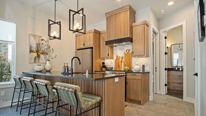 A modern kitchen with wooden cabinetry, a central island with bar stools, pendant lighting, and a view of the adjacent living area.