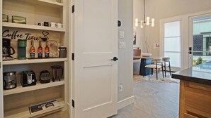 A modern kitchen with open shelves displaying kitchenware and appliances, a dining area with a table and chairs, and a view of the outside through a glass door.