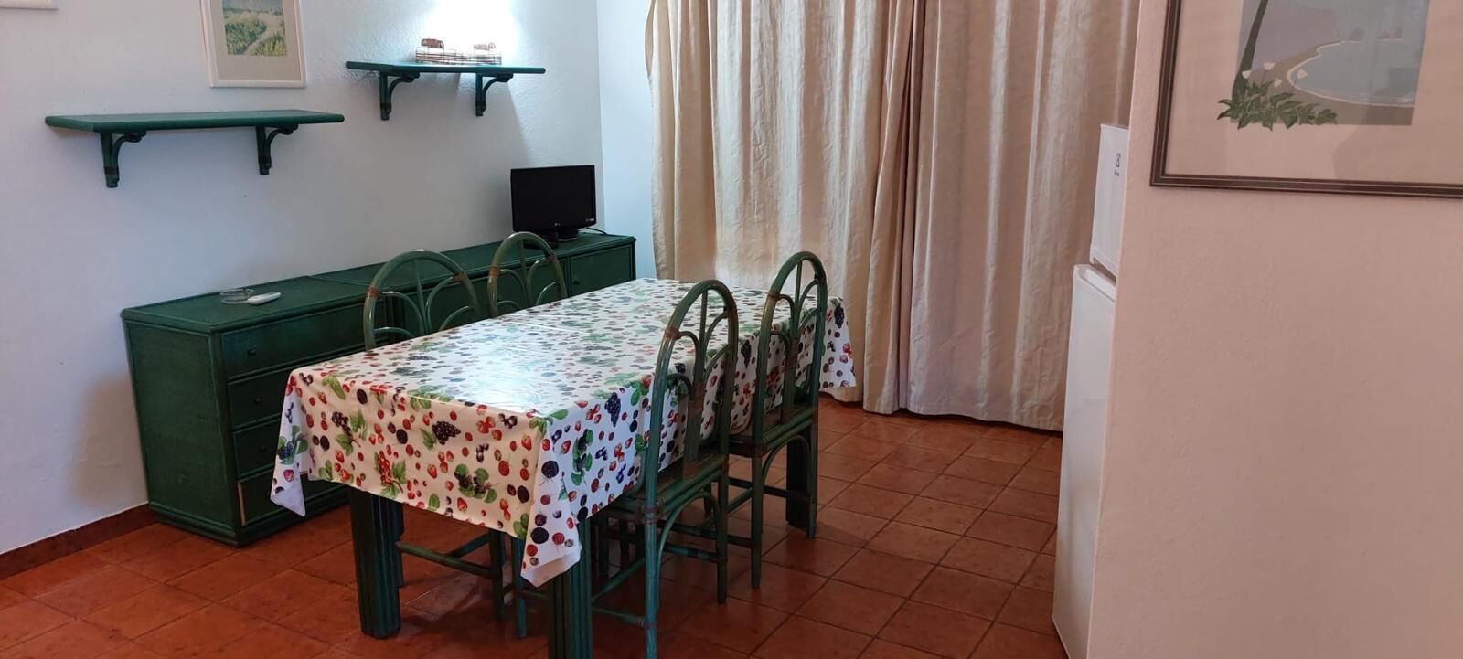A dining area with a table covered in a fruit-patterned tablecloth, green chairs, a green cabinet, and a television on a stand.