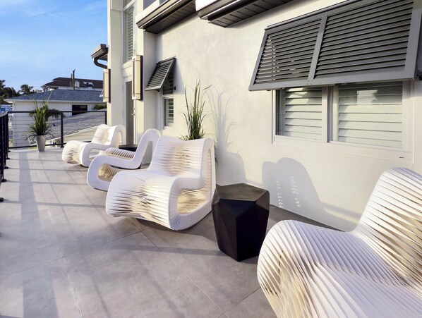 Un espacio exterior moderno con asientos de mimbre blanco y una mesa negra, junto a un edificio con ventanas y persianas grandes.